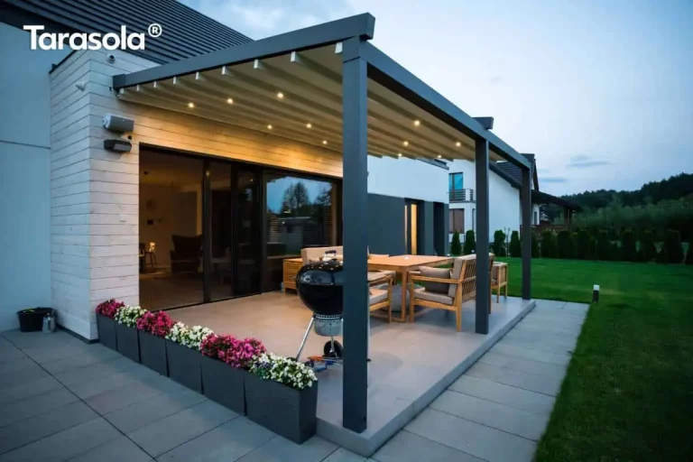 Nowoczesna, przyścienna pergola tarasowa Elegancy Tarasola zamontowana nad betonowym tarasem przy jednorodzinnym budynku o jasnej elewacji. Konstrukcja wykonana jest z ekstrudowanych profili aluminiowych w kolorze antracytowym, wsparta na dwóch masywnych przednich słupach. Zadaszenie stanowi ruchoma, biała tkanina ułożona w charakterystyczne fale, która jest w pełni rozłożona. W poprzecznych profilach podtrzymujących materiał zintegrowano punktowe oświetlenie LED, emitujące ciepłe światło. Pod pergolą znajduje się strefa wypoczynkowa z drewnianymi meblami ogrodowymi, szarymi poduszkami oraz czarnym grillem węglowym. Taras otoczony jest niskimi, ciemnoszarymi donicami z kolorowymi kwiatami, a w tle widoczny jest zadbany trawnik oraz sąsiednia zabudowa w nowoczesnym stylu.