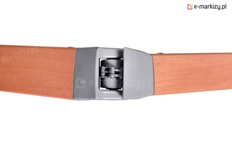 Zbliżenie na aluminiowy przegub ramienia markizy w okleinie drewnopodobnej. Centralnym punktem zdjęcia jest mechanizm łączący dwa profile ramienia, wykonany z szarego odlewu. Wewnątrz przegubu widoczny jest stalowy łańcuch wielorzędowy (łańcuch Fleyera), który odpowiada za napięcie ramienia podczas rozkładania i składania osłony. Profile ramienia mają prostokątny przekrój i są wykończone gładką powłoką o strukturze i kolorze jasnego drewna z wyraźnym rysunkiem słojów. Konstrukcja jest przedstawiona na białym, neutralnym tle w rzucie z góry lub z boku, ukazując precyzyjne spasowanie elementów metalowych z profilami nośnymi.