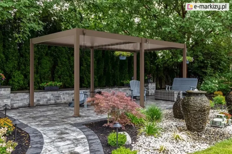 Duża wolnostojąca pergola lamelowa Marlo w kolorze taupe, zamontowana w starannie zaaranżowanym ogrodzie na podłożu z ciemnoszarej kostki brukowej. Konstrukcja składa się z dwóch modułów zadaszenia wspartych na sześciu aluminiowych słupach o kwadratowym przekroju. Ruchome lamele dachowe są ustawione w pozycji zamkniętej, tworząc pełną osłonę przed słońcem. Przy słupach widoczne są mechanizmy korbowe służące do ręcznej regulacji dachu. W strefie pod pergolą znajduje się szary fotel ogrodowy oraz dopasowana sofa wykonana z technorattanu. Taras jest otoczony niskim murkiem z jasnoszarych kamiennych bloczków, za którym rozciąga się gęsty żywopłot z tui oraz wysokie drzewa liściaste. Na pierwszym planie widoczna jest nowoczesna rabata z jasnym żwirem, dekoracyjnymi głazami, dużymi ozdobnymi wazami oraz czerwonolistnym klonem palmowym. Wzdłuż ścieżki i na rabatach rozmieszczono niskie, czarne lampy ogrodowe.
