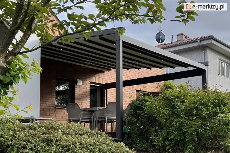 Pergola tarasowa o aluminiowej konstrukcji w kolorze ciemnego antracytu, zamontowana do ściany budynku wykonanej z czerwonej cegły klinkierowej. Zadaszenie pergoli tworzy jasny, kremowy materiał ułożony w regularne pasy, zamocowane na poprzecznych profilach, w których umieszczono punktowe oświetlenie LED. Konstrukcja wspiera się na grubym, pionowym słupie o kwadratowym przekroju, a boczna część została uzupełniona o poziomą belkę stabilizującą. Pod zadaszeniem, na drewnianym tarasie, widoczne są krzesła ogrodowe z szarej plecionki ustawione przy stole. Całość otoczona jest gęstą roślinnością ogrodową, w tym wysokimi zielonymi krzewami, które częściowo zasłaniają dolną część konstrukcji. W górnej części kadru widoczne są gałęzie liściastego drzewa z zielonymi liśćmi, a w tle zarys sąsiedniego budynku o szarej elewacji z anteną satelitarną na dachu pod zachmurzonym niebem.