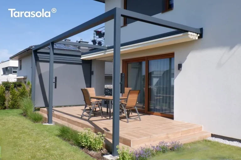Zdjęcie przedstawia aluminiową pergolę tarasową Tarasola Flat o konstrukcji przyściennej w kolorze ciemnego antracytu, zamontowaną przy nowoczesnym domu jednorodzinnym. Zadaszenie tkaninowe w kolorze kremowym jest w tym momencie całkowicie zwinięte pod systemowy daszek ochronny, co odsłania pełną ramę konstrukcji oraz przestrzeń tarasu. Na jednym z boków pergoli zainstalowany jest pionowy refleksol Zip z tkaniną typu screen w kolorze szarym, która jest w pełni opuszczona, zapewniając osłonę boczną. Taras został wyłożony płytkami w ciepłym odcieniu brązu i wyposażony w drewniany stół oraz cztery krzesła z tekstylnym obiciem. Na słupach konstrukcyjnych widoczne są zamontowane akcesoria, prawdopodobnie kinkiety lub głośniki. Otoczenie stanowi zadbany trawnik z niskimi nasadzeniami lawendy i traw ozdobnych wzdłuż krawędzi podestu. Elewacja budynku jest jasna, z dużymi przesuwnymi drzwiami tarasowymi w drewnianej ramie, a w tle widoczne są sąsiednie zabudowania z panelami fotowoltaicznymi na dachu.