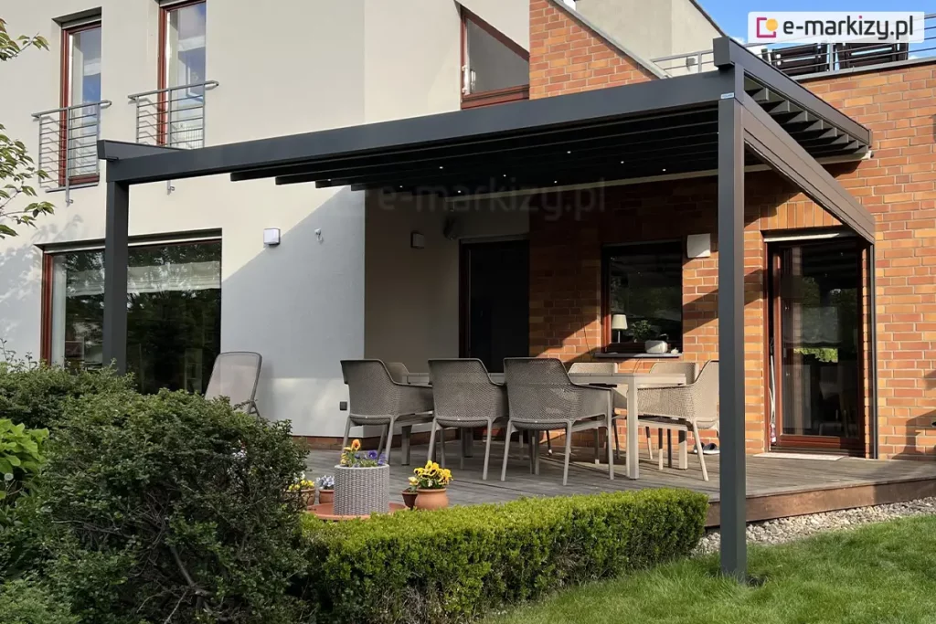 Aluminiowa Pergola tarasowa Square o konstrukcji w kolorze ciemnego antracytu, zamontowana do elewacji budynku, która łączy tynk w kolorze złamanej bieli z fragmentami wykonanymi z czerwonej cegły klinkierowej. Zadaszenie pergoli wykonane jest z tkaniny w ciemnym kolorze, pod którą zainstalowano punktowe oświetlenie LED, rozmieszczone regularnie na profilach poprzecznych. Konstrukcja opiera się na dwóch pionowych słupach o przekroju kwadratowym, a z prawej strony posiada dodatkową belkę usztywniającą, do której zamocowana została zwinięta osłona boczna refleksol Zip l Zip U. Na tarasie wyłożonym deskami kompozytowymi w odcieniu naturalnego drewna ustawiono duży stół ogrodowy oraz sześć dopasowanych krzeseł z technorattanu w kolorze beżowym. W tle widoczne są duże przeszklenia drzwi tarasowych oraz okna z białymi ramami, a na piętrze balkon z metalową, ażurową balustradą. Przed tarasem znajduje się zadbany ogród z zielonym trawnikiem, niskim, formowanym żywopłotem oraz donicami z kwitnącymi kwiatami w kolorze żółtym i fioletowym. Całość oświetlona jest naturalnym światłem dziennym, tworząc funkcjonalną przestrzeń do wypoczynku na świeżym powietrzu.