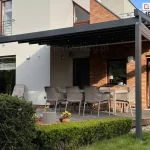 Aluminiowa Pergola tarasowa Square o konstrukcji w kolorze ciemnego antracytu, zamontowana do elewacji budynku, która łączy tynk w kolorze złamanej bieli z fragmentami wykonanymi z czerwonej cegły klinkierowej. Zadaszenie pergoli wykonane jest z tkaniny w ciemnym kolorze, pod którą zainstalowano punktowe oświetlenie LED, rozmieszczone regularnie na profilach poprzecznych. Konstrukcja opiera się na dwóch pionowych słupach o przekroju kwadratowym, a z prawej strony posiada dodatkową belkę usztywniającą, do której zamocowana została zwinięta osłona boczna refleksol Zip l Zip U. Na tarasie wyłożonym deskami kompozytowymi w odcieniu naturalnego drewna ustawiono duży stół ogrodowy oraz sześć dopasowanych krzeseł z technorattanu w kolorze beżowym. W tle widoczne są duże przeszklenia drzwi tarasowych oraz okna z białymi ramami, a na piętrze balkon z metalową, ażurową balustradą. Przed tarasem znajduje się zadbany ogród z zielonym trawnikiem, niskim, formowanym żywopłotem oraz donicami z kwitnącymi kwiatami w kolorze żółtym i fioletowym. Całość oświetlona jest naturalnym światłem dziennym, tworząc funkcjonalną przestrzeń do wypoczynku na świeżym powietrzu.