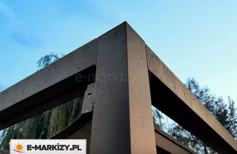 Zbliżenie narożnika pergoli Dragon, lekko zroszonego porannym deszczem. Krople deszczu wyraźnie widoczne na tle antracytowej konstrukcji. Ponad aluminiowym narożnikiem pergoli widoczne niebieskie niebo.