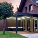 Aluminiowa pergola sb350 Aluprof z podświetleniem w obwodzie dachu na tarasie przy ceglanym domu wieczorową porą. Jasne pióra lameli kontrastują z grafitową konstrukcją