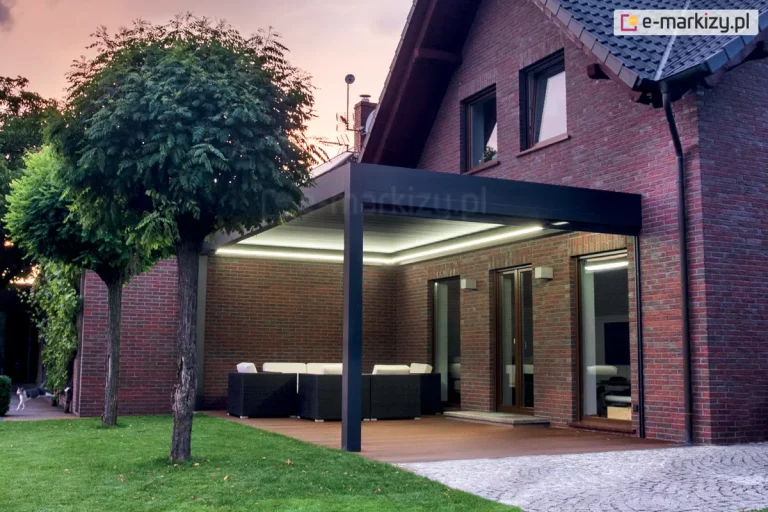 Aluminiowa pergola sb350 Aluprof z podświetleniem w obwodzie dachu na tarasie przy ceglanym domu wieczorową porą. Jasne pióra lameli kontrastują z grafitową konstrukcją