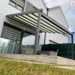 Wizualizacja nowoczesnej pergoli tarasowej o grafitowej, aluminiowej konstrukcji przyściennej, uchwycona w ujęciu od dołu, z perspektywy trawnika. Pergola tarasowa przyścienna Aura składa się z kremowego materiału technicznego rozpiętego na równoległych profilach poprzecznych, które są w pełni wysunięte, tworząc lekką, schodkową strukturę. Konstrukcja wspiera się na dwóch solidnych, kwadratowych słupach bocznych o antracytowym wykończeniu. Pod pergolą, na podwyższonym tarasie wyłożonym jasnobeżowymi płytami wielkoformatowymi, widoczny jest zestaw mebli ogrodowych wykonanych z szarego technorattanu. Tło stanowi elewacja nowoczesnego budynku o białym tynku z dużym przeszkleniem drzwi tarasowych, w których odbija się otoczenie. Po prawej stronie taras jest odgrodzony od sąsiedniej posesji za pomocą ciemnozielonej siatki cieniującej zamontowanej na ogrodzeniu. Niebo nad budynkiem jest jasne, z dynamicznie ułożonymi białymi chmurami, co podkreśla nowoczesną architekturę i estetykę instalacji.