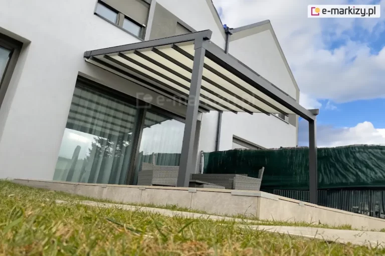 Wizualizacja nowoczesnej pergoli tarasowej o grafitowej, aluminiowej konstrukcji przyściennej, uchwycona w ujęciu od dołu, z perspektywy trawnika. Pergola tarasowa przyścienna Aura składa się z kremowego materiału technicznego rozpiętego na równoległych profilach poprzecznych, które są w pełni wysunięte, tworząc lekką, schodkową strukturę. Konstrukcja wspiera się na dwóch solidnych, kwadratowych słupach bocznych o antracytowym wykończeniu. Pod pergolą, na podwyższonym tarasie wyłożonym jasnobeżowymi płytami wielkoformatowymi, widoczny jest zestaw mebli ogrodowych wykonanych z szarego technorattanu. Tło stanowi elewacja nowoczesnego budynku o białym tynku z dużym przeszkleniem drzwi tarasowych, w których odbija się otoczenie. Po prawej stronie taras jest odgrodzony od sąsiedniej posesji za pomocą ciemnozielonej siatki cieniującej zamontowanej na ogrodzeniu. Niebo nad budynkiem jest jasne, z dynamicznie ułożonymi białymi chmurami, co podkreśla nowoczesną architekturę i estetykę instalacji.