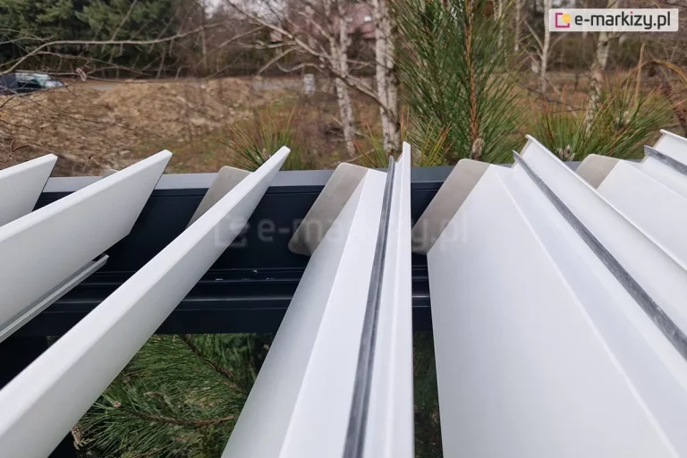 Białe aluminiowe lamele pergoli Aluprof z antracytową konstrukcją, wyposażone w czarne gumowe uszczelki wzdłuż powierzchni styku paneli. W tle widoczne drzewa .