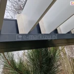 Zbliżenie na mechanizm mocowania lameli w aluminiowej pergoli tarasowej Sb350 Aluprof. Zdjęcie przedstawia punkt styku białych, ruchomych lameli z ciemnoantracytową ramą konstrukcyjną. Widoczne są srebrne, metalowe zakończenia lameli, które są precyzyjnie osadzone w prowadnicy ramy, umożliwiając ich obrót. Lamele są ustawione w pozycji otwartej. W dolnej części kadru widoczne są igły zielonej sosny, a w tle rozmyty zarys bezlistnych drzew liściastych na tle jasnego nieba. Fotografia detalu ukazuje wysoką jakość wykończenia i dopasowania elementów konstrukcyjnych pergoli bioklimatycznej.
