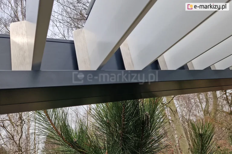 Zbliżenie na mechanizm mocowania lameli w aluminiowej pergoli tarasowej Sb350 Aluprof. Zdjęcie przedstawia punkt styku białych, ruchomych lameli z ciemnoantracytową ramą konstrukcyjną. Widoczne są srebrne, metalowe zakończenia lameli, które są precyzyjnie osadzone w prowadnicy ramy, umożliwiając ich obrót. Lamele są ustawione w pozycji otwartej. W dolnej części kadru widoczne są igły zielonej sosny, a w tle rozmyty zarys bezlistnych drzew liściastych na tle jasnego nieba. Fotografia detalu ukazuje wysoką jakość wykończenia i dopasowania elementów konstrukcyjnych pergoli bioklimatycznej.