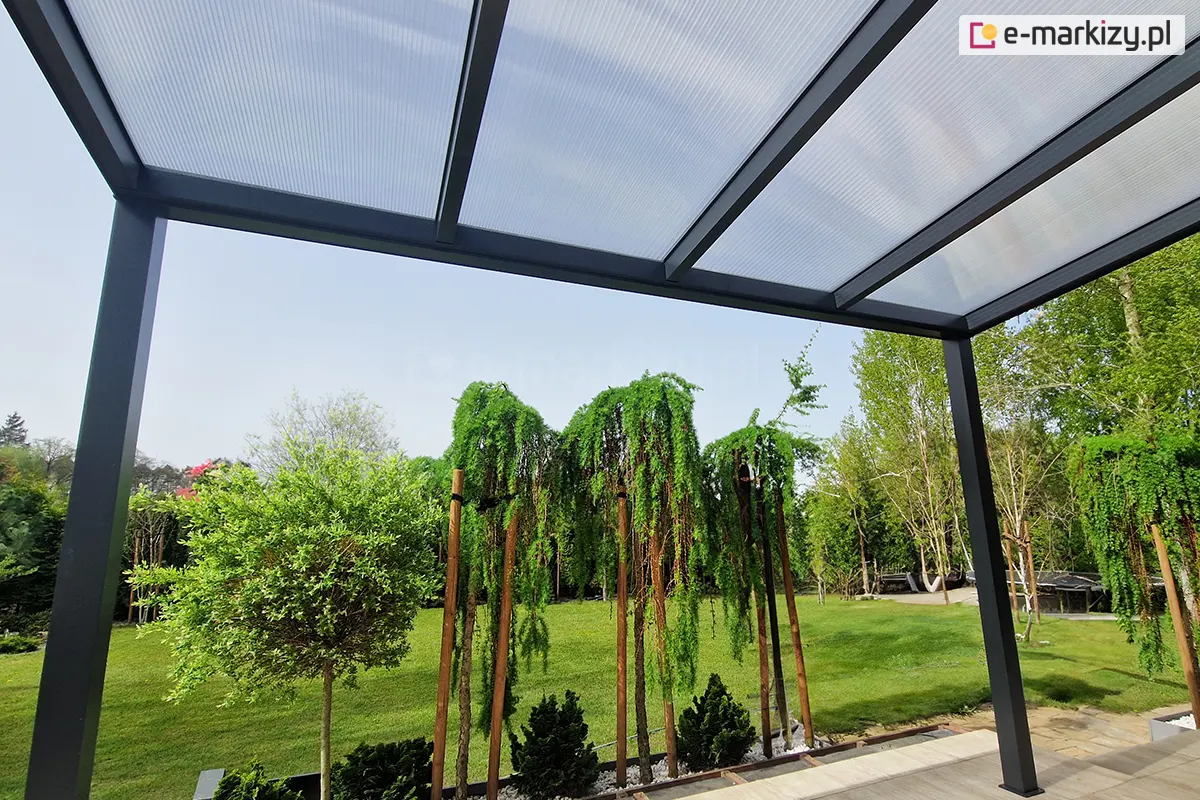 Zdjęcie wykonane z perspektywy żabiej, ukazujące od spodu konstrukcję aluminiowego zadaszenia w kolorze antracytowym. Widoczne są równolegle rozmieszczone krokwie podtrzymujące płyty poliwęglanu komorowego, przez które przebija rozproszone światło dzienne. Konstrukcja opiera się na dwóch skrajnych słupach, tworząc ramę dla widoku na ogród. W tle widać zielony trawnik oraz ozdobne drzewa iglaste o płaczącym pokroju, posadzone w pasie białego żwiru.