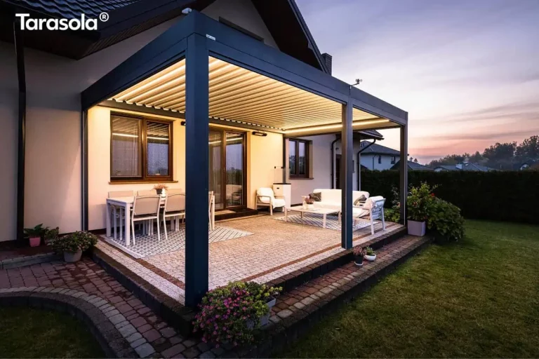 Nowoczesna, przyścienna pergola bioklimatyczna essential Tarasola w kolorze ciemnoantracytowym, zamontowana nad tarasem wykonanym z drobnej kostki brukowej. Konstrukcja posiada dach z ruchomymi lamelami ustawionymi pod kątem oraz zintegrowane oświetlenie LED w formie listew biegnących wzdłuż wewnętrznej krawędzi ramy, które emituje ciepłe światło. Pod zadaszeniem zaaranżowano strefę jadalnianą z białym stołem i krzesłami oraz strefę wypoczynkową z białą sofą i stolikiem kawowym ustawionym na dekoracyjnym dywanie. Pergola przylega do jasnej elewacji domu jednorodzinnego z brązową stolarką okienną, a cała instalacja jest posadowiona na podwyższeniu z ciemnym obrzeżem. Zdjęcie wykonane o zmierzchu ukazuje nastrojową atmosferę ogrodu z widocznym w tle trawnikiem, ciemnym żywopłotem oraz subtelnym, wieczornym niebem w odcieniach fioletu i pomarańczu.