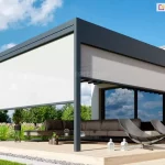 Zdjęcie przedstawia nowoczesną pergolę tarasową o konstrukcji przyściennej, wykonaną z aluminiowych profili w kolorze ciemnoszarym (antracytowym). Zadaszenie ma formę płaską, a dach został wykonany z wodoodpornej tkaniny układającej się w charakterystyczne fale. Konstrukcja jest zamontowana na drewnianym podeście tarasowym otoczonym pasem szarego żwiru i trawnikiem. Dwa boki pergoli są częściowo osłonięte pionowymi roletami zewnętrznymi typu screen w kolorze jasnokremowym lub białym, które służą do ochrony przed słońcem i wiatrem. Frontowa roleta jest opuszczona do połowy wysokości, natomiast boczna jest podciągnięta wyżej. Pod zadaszeniem znajduje się zestaw mebli ogrodowych, w tym narożna sofa z ciemnoszarymi poduchami oraz dwa nowoczesne fotele o ażurowej konstrukcji. Na tarasie widoczne są również elementy dekoracyjne: lampiony, donice z roślinami oraz ozdobne trawy ogrodowe w tle. Budynek, do którego przylega taras, posiada elewację wykończoną białą farbą oraz fragmenty z cegły klinkierowej w kolorze pomarańczowo-brązowym, z dużymi przeszkleniami odbijającymi niebo.