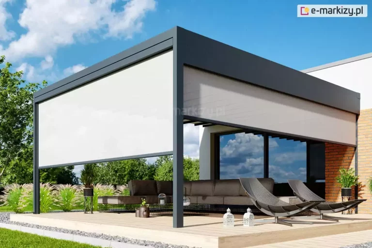 Zdjęcie przedstawia nowoczesną pergolę tarasową o konstrukcji przyściennej, wykonaną z aluminiowych profili w kolorze ciemnoszarym (antracytowym). Zadaszenie ma formę płaską, a dach został wykonany z wodoodpornej tkaniny układającej się w charakterystyczne fale. Konstrukcja jest zamontowana na drewnianym podeście tarasowym otoczonym pasem szarego żwiru i trawnikiem. Dwa boki pergoli są częściowo osłonięte pionowymi roletami zewnętrznymi typu screen w kolorze jasnokremowym lub białym, które służą do ochrony przed słońcem i wiatrem. Frontowa roleta jest opuszczona do połowy wysokości, natomiast boczna jest podciągnięta wyżej. Pod zadaszeniem znajduje się zestaw mebli ogrodowych, w tym narożna sofa z ciemnoszarymi poduchami oraz dwa nowoczesne fotele o ażurowej konstrukcji. Na tarasie widoczne są również elementy dekoracyjne: lampiony, donice z roślinami oraz ozdobne trawy ogrodowe w tle. Budynek, do którego przylega taras, posiada elewację wykończoną białą farbą oraz fragmenty z cegły klinkierowej w kolorze pomarańczowo-brązowym, z dużymi przeszkleniami odbijającymi niebo.