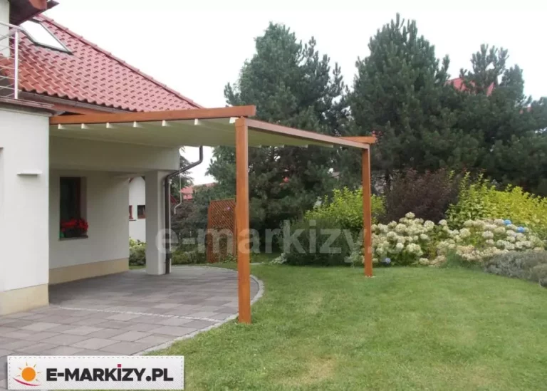 Automatyczne zadaszenie tarasu w formie nowoczesnej pergoli Luxo o konstrukcji wykonanej z aluminium pokrytego lakierem w kolorze naturalnego drewna w ciepłym odcieniu brązu. Zadaszenie jest zamontowane jednostronnie do białej elewacji piętrowego budynku mieszkalnego, a z drugiej strony oparte na dwóch solidnych pionowych słupach o przekroju kwadratowym. Pod poszyciem widoczne są poprzeczne profile nośne, które podtrzymują jasną, kremową tkaninę chroniącą przed słońcem i deszczem. Pergola osłania taras wyłożony szarą kostką brukową o prostokątnym kształcie. W tle znajduje się zadbany ogród z zielonym trawnikiem, gęstymi krzewami ozdobnymi, białymi hortensjami oraz wysokimi drzewami iglastymi. Nad konstrukcją widoczny jest fragment dachu budynku pokrytego czerwoną dachówką ceramiczną z oknem połaciowym. Całość przedstawia funkcjonalną zabudowę zewnętrznej strefy wypoczynkowej zintegrowaną z architekturą domu i otaczającą roślinnością.
