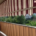Zdjęcie przedstawia produkt, którym jest markiza balkonowa poręczowa zamontowana na balkonie w budynku mieszkalnym. Markiza wykonana jest z materiału w pionowe pasy o kolorach bordowym, białym i jasnobrązowym, a jej dolna krawędź posiada charakterystyczne ozdobne wykończenie w formie fali, obszyte czerwoną lamówką. Tkanina jest rozciągnięta na metalowej konstrukcji, która pozwala na regulację kąta nachylenia osłony. Markiza balkonowa poręczowa jest tu ustawiona w pozycji częściowo rozwiniętej, tworząc cień na powierzchni balkonu i chroniąc wnętrze przed bezpośrednim nasłonecznieniem. Poniżej materiału markizy widoczna jest balustrada balkonowa, która została dodatkowo osłonięta pionową przesłoną w kolorze beżowym, pasującą kolorystycznie do całości aranżacji. Na balkonie, po lewej stronie kadru, znajduje się czarny grill ogrodowy na kółkach. W tle za barierką rozpościera się widok na gęstą, zieloną roślinność ogrodową oraz fragmenty sąsiedniej zabudowy jednorodzinnej pod bezchmurnym, jasnym niebem. Całość kompozycji ukazuje praktyczne zastosowanie markizy balkonowej poręczowej jako skutecznego rozwiązania przeciwsłonecznego, które jednocześnie zapewnia prywatność użytkownikom tarasu.