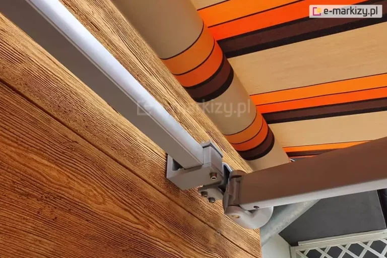 Zbliżenie na mechanizm montażowy oraz konstrukcję markizy tarasowej Allegro zamocowanej do elewacji budynku wykończonej deskami o wyraźnej strukturze drewna w ciepłym brązowym odcieniu. Na pierwszym planie widoczny jest solidny aluminiowy profil kwadratowy w kolorze jasnoszarym, który stanowi główną belkę nośną konstrukcji. Belka ta jest osadzona w masywnym uchwycie ściennym wykonanym z odlewu aluminium, przykręconym do drewnianej ściany za pomocą stalowych śrub. Do belki nośnej przymocowany jest przegub ramienia składanego, który umożliwia płynne rozwijanie i zwijanie poszycia. Ramię markizy ma opływowy kształt i jest dopasowane kolorystycznie do pozostałych elementów stelaża. W górnej części kadru widoczna jest rura nawojowa, na którą nawinięty jest materiał markizowy. Tkanina posiada wzór w szerokie pasy w kolorystyce pomarańczowej, beżowej oraz brązowej. Materiał charakteryzuje się gęstym splotem, typowym dla tkanin akrylowych stosowanych w markizach tarasowych Allegro, co zapewnia ochronę przed słońcem. Całość konstrukcji prezentuje się nowocześnie i stabilnie, ukazując detale techniczne połączeń mechanicznych oraz estetykę wykończenia detali.