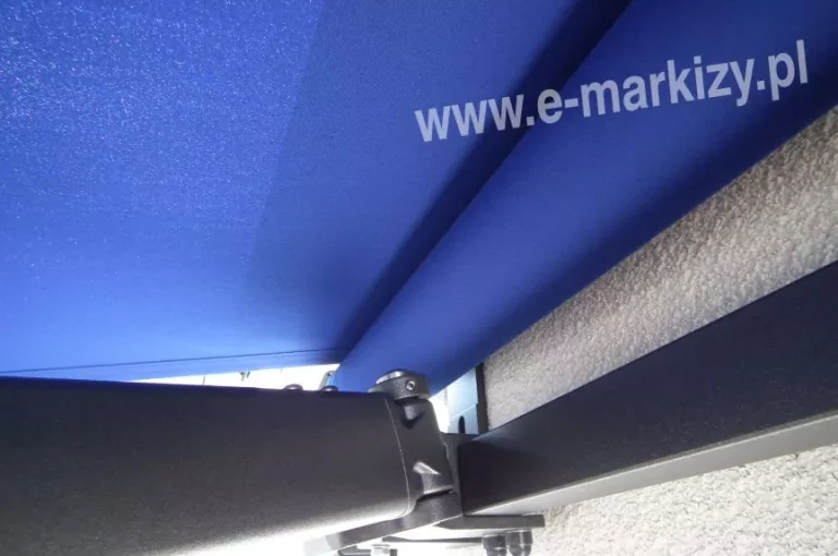 Zdjęcie przedstawia zbliżenie na detale techniczne i wykończenie konstrukcji, jaką posiada markiza Australia Aluprof. Na pierwszym planie widoczny jest solidny, aluminiowy profil nośny oraz przegub ramienia w kolorze ciemnego grafitu o matowej, lekko chropowatej strukturze. Elementy konstrukcyjne są połączone za pomocą precyzyjnych śrub imbusowych, co podkreśla techniczny charakter produktu. Powyżej ramion rozpięte jest poszycie wykonane z wysokiej jakości tkaniny akrylowej w intensywnym, chabrowym odcieniu niebieskiego. Materiał jest nawinięty na rurę nawojową, co jest charakterystyczne dla modelu markiza Australia Aluprof w wersji bezkasetowej. Tkanina wykazuje gęsty splot, a światło padające od góry uwydatnia jej fakturę oraz nasycenie koloru. Wąski kadr skupia się na punkcie mocowania ramienia do belki kwadratowej, pokazując stabilność i masywność mechanizmu. Tło dla konstrukcji stanowi jasna, chropowata elewacja budynku o strukturze tynku typu baranek. Perspektywa zdjęcia od dołu pozwala dokładnie przyjrzeć się mechanice działania ramion przegubowych oraz sposobowi, w jaki tkanina układa się na rolce.
