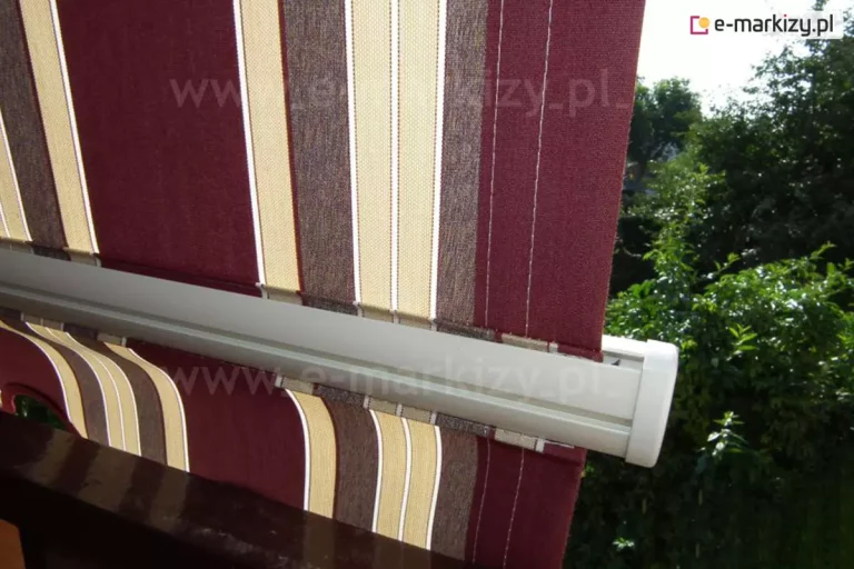 Belka przednia markizy balkonowej-poręczowej w kolorze białym wyraźnie szersza niż zamocowane do niej poszycie oraz falbana w brodowe, żółte, białe i brązowe pasy z widocznymi od wewnątrz przeszyciami. W tle widoczne drzewa o zielonych, oświetlonych słońcem liściach.