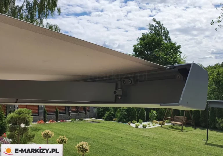 Aluminiowa belka przednia nowoczesnej markizy elektrycznej Moderno sfotografowana w zbliżeniu pod lekkim kątem od dołu. Zdjęcie o charakterze technicznym przedstawia prawy skrajny narożnik profilu czołowego oraz połączone z nim ramię przegubowe w pozycji wysuniętej. Konstrukcja profilu charakteryzuje się nowoczesną, minimalistyczną formą o lekko zaokrąglonym kształcie, zamkniętą z boku płaską, dopasowaną zaślepką z tworzywa sztucznego. Całość stelaża wykończona jest matową powłoką lakierniczą w kolorze głębokiego grafitu lub ciemnego antracytu. Wewnątrz profilu widoczne są śruby montażowe imbusowe mocujące przegub ramienia nośnego. Nad aluminiową konstrukcją rozciąga się równomiernie napięta tkanina poszycia markizowego w gładkim, jednolitym odcieniu jasnego beżu lub kremu. Dolna krawędź materiału wchodzi bezpośrednio w specjalny rowek montażowy belki frontowej, zapewniając estetyczne i bezszwowe wykończenie bez tradycyjnej falbany. W tle, poniżej linii markizy, widoczna jest przestrzenna aranżacja przydomowego ogrodu z zadbanym, zielonym trawnikiem, ozdobnymi krzewami formowanymi w kule na pniu oraz niskimi nasadzeniami w pasie grysowym. Za trawnikiem wznosi się nowoczesne ogrodzenie posesji z poziomymi, ciemnobrązowymi przęsłami drewnianymi lub kompozytowymi, a po prawej stronie stoi drewniana huśtawka ogrodowa. Górną część tła wypełnia błękitne niebo pokryte gęstymi, białymi chmurami kłębiastymi oraz wierzchołki zielonych drzew liściastych i brzóz.
