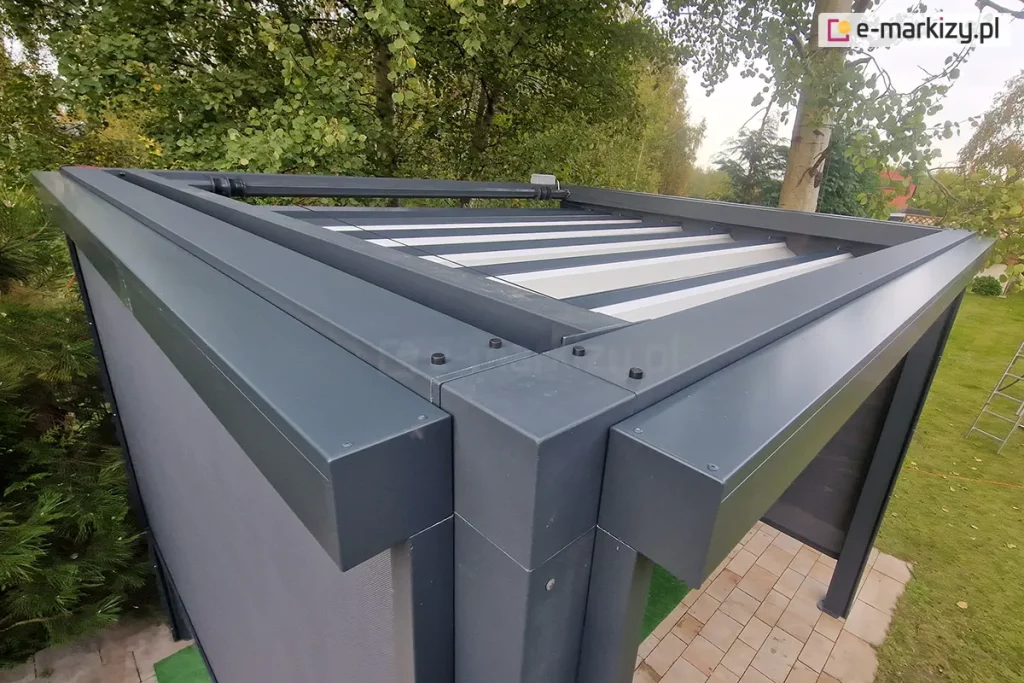 Dwie skrzynki refleksoli zamocowane do ścian bocznych pergoli Tako. Po lewej rozsunięta tkanina rolety w kolorze grafitowym, po prawej widoczne pod pergolą podłoże z kostki brukowej. Dookoła pergoli wiele drzew liściastych.