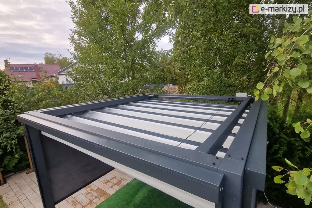Widok z góry na konstrukcję pergoli tarasowej Tako w kolorze antracytowym, prezentujący mechanizm zadaszenia tkaninowego. Na pierwszym planie widoczna jest masywna górna rama wykonana z profili aluminiowych o matowym wykończeniu. Wewnątrz ramy znajduje się całkowicie rozsunięty system zadaszenia rzymskiego w kolorze jasnokremowym. Widoczne są poprzeczne profile usztywniające tkaninę oraz stalowe linki prowadzące, które zapewniają stabilność i precyzyjne zwijanie materiału. Z trzech stron pergoli zamontowane są rolety boczne - refleksole Ziiip w kolorze ciemnoszarym. Pergola jest ustawiona na podłożu z jasnej kostki brukowej, z wydzieloną strefą zielonej sztucznej trawy. Tło zdjęcia stanowią gęste korony drzew liściastych z zielonymi i żółknącymi liśćmi, a w oddali widoczny jest fragment dachu sąsiedniego budynku mieszkalnego z zainstalowanymi panelami fotowoltaicznymi. Niebo jest zachmurzone, co zapewnia miękkie, rozproszone oświetlenie całej konstrukcji.