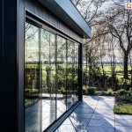 Zdjęcie przedstawia nowoczesną, bezramową zabudowę tarasu wykonaną z ruchomych paneli ze szkła hartowanego, zamontowanych w konstrukcji o barwie antracytowej. Całość przeszklenia składa się z kilku wysokich, pionowych tafli ulokowanych w dolnej szynie prowadzącej. Konstrukcja zadaszenia posiada wbudowane w podbitkę punktowe oświetlenie LED, a boczna ściana budynku wykończona jest pionowymi panelami elewacyjnymi w ciemnym kolorze. Przed zabudową rozciąga się taras wyłożony dużymi, kwadratowymi płytami w odcieniu szarości, przechodzący w zadbany trawnik z niskimi nasadzeniami roślinnymi. W gładkich, przezroczystych powierzchniach szklanych odbija się otaczający krajobraz, w tym wysokie, bezlistne drzewa oraz rozległe, zielone pola pod jasnym niebem, co tworzy efekt lustrzanego odbicia natury na elewacji obiektu.