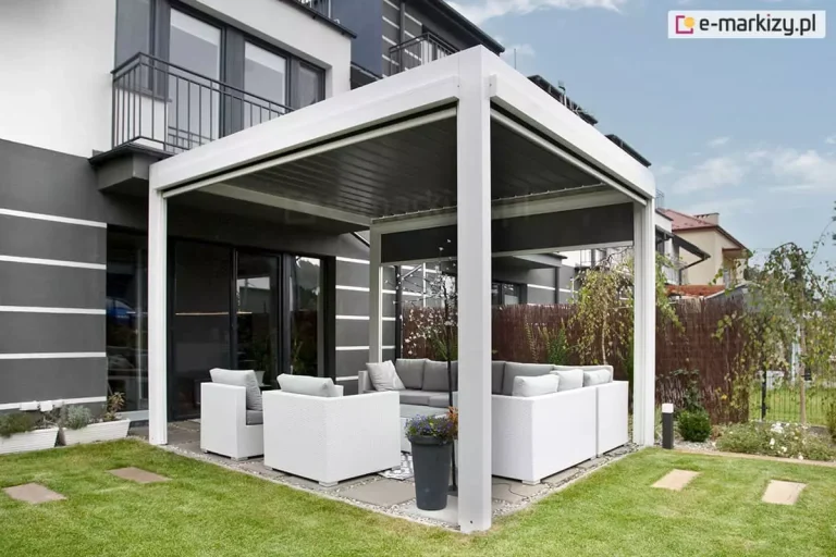Pergola lamelowa wolnostojąca Sb400 Aluprof w kolorze białym z dodatkowymi refleksolami na trzech ścianach ustawiona nad niewielkim tarasem z jasnymi meblami w sąsiedztwie zielonego trawnika tuż przy wyjściu na taras z piętrowego domku.