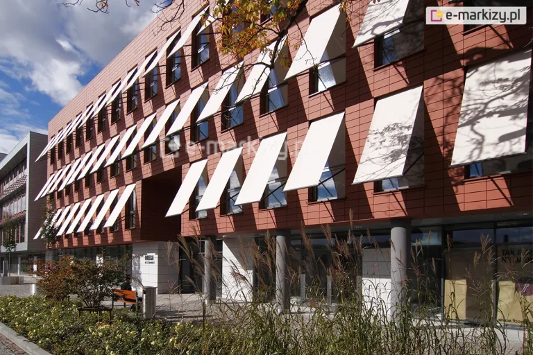 Elewacja nowoczesnego budynku biurowego, wykończona ceramicznymi płytami w kolorze ceglastym, z rzędami pionowych okien wyposażonych w system zewnętrznych osłon przeciwsłonecznych. Na trzech kondygnacjach obiektu zamontowano liczne markizy sklepowe Italia Aluprof z tkaniną akrylową w jasnym, niemal białym kolorze, które są wysunięte pod kątem, tworząc powtarzalny, geometryczny wzór na fasadzie. Parter budynku jest przeszklony i cofnięty względem lica wyższych pięter, wsparty na szarych, cylindrycznych słupach konstrukcyjnych. Przed budynkiem znajduje się zagospodarowany teren z niskimi krzewami, ozdobnymi trawami oraz elementami małej architektury, takimi jak metalowy kosz na śmieci i drewniana ławka. W prawej części kadru na jasnej tkaninie markiz widoczne są cienie rzucane przez gałęzie pobliskiego drzewa. Niebo jest częściowo zachmurzone, a naturalne oświetlenie podkreśla kontrast między ciepłym odcieniem elewacji a jasnymi płaszczyznami rozwiniętych osłon.