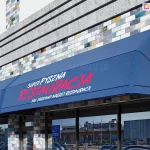 Umieszczone na ścianie z kolorowych cegieł nad dużymi oknami witrynowymi oraz wejściem do budynku trzy markizy koszowe proste o różnej szerokości, wyposażone w niebieskie poszycia oraz proste falbany z różnymi nadrukami na polach nad falbaną tworzą spójną reklamę restauracji.