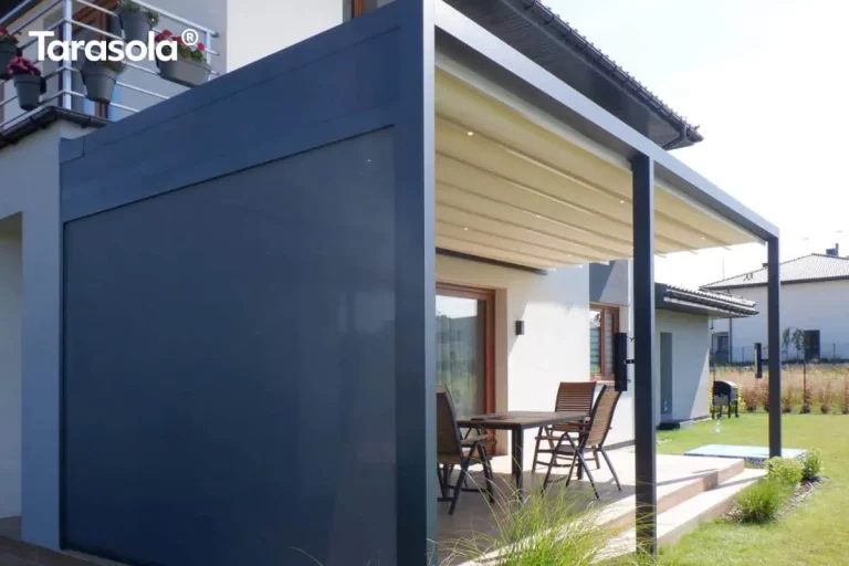 Zdjęcie prezentuje pergolę przyścienną Tarasola Flat o antracytowej konstrukcji aluminiowej, zamontowaną przy białej elewacji budynku jednorodzinnego. Konstrukcja wspiera się na trzech równomiernie rozłożonych słupach podporowych o kwadratowym przekroju. Model ten posiada dach tkaninowy w kolorze kremowym, który jest w pełni rozwinięty i wyposażony w zintegrowane, punktowe oświetlenie LED rozmieszczone na profilach poprzecznych. Charakterystycznym elementem tej realizacji jest boczna osłona typu screen (zip) w kolorze ciemnoszarym, która jest całkowicie opuszczona, zamykając jeden z boków pergoli i zapewniając ochronę przed wiatrem oraz słońcem. Pod zadaszeniem, na tarasie wyłożonym jasnobrązowymi płytkami, ustawiono drewniany stół z krzesłami. W tle widoczny jest zielony trawnik, niskie nasadzenia ozdobne oraz sąsiednie domy jednorodzinne pod czystym, błękitnym niebem.