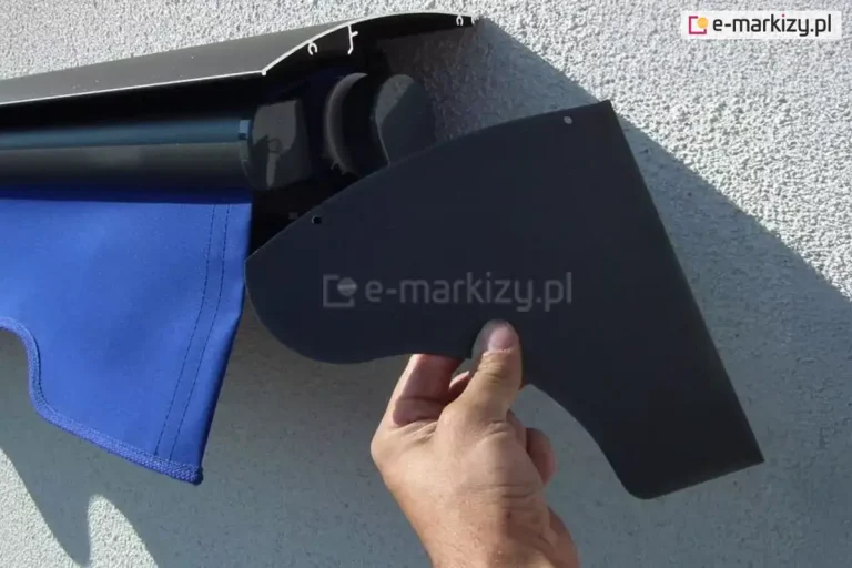Ludzka ręka prezentująca boczną zaślepkę daszka do markiz 22,5 cm z widoczną w tle markizą Aluprof z daszkiem w kolorze grafitowym i falbaną falowaną w chabrowym kolorze, zamocowaną na ścianie z białym tynkiem strukturalnym