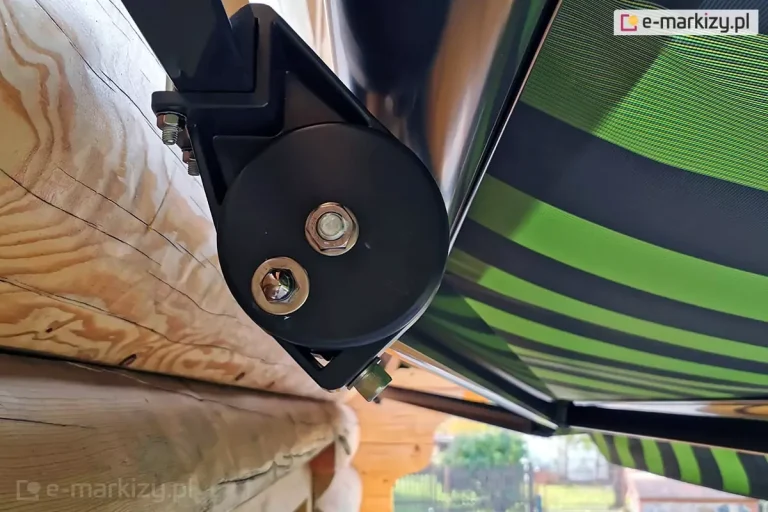 Zbliżenie na boczny mechanizm mocujący markizę Adagio zainstalowaną na ścianie wykonanej z naturalnych bali drewnianych. Konstrukcja uchwytu jest wykonana z metalu malowanego proszkowo na kolor ciemnografitowy lub czarny. Widoczny jest boczny profil wspornika, który jest solidnie przykręcony do drewna. Centralnym elementem zdjęcia jest okrągła, czarna osłona mechanizmu przegubowego, na której znajdują się dwie duże śruby montażowe z podkładkami. Górna śruba posiada sześciokątny łeb, natomiast dolna jest zakończona ozdobną nakrętką kołpakową. Nad mechanizmem przebiega rura nawojowa markizy Adagio, a z boku widoczna jest krawędź poszycia materiałowego w pasy o różnych odcieniach zieleni oraz czerni. Materiał jest napięty i rozłożony, co pozwala dostrzec splot tkaniny. W tle, pod tkaniną, zarysowują się ramiona przegubowe wspierające konstrukcję oraz fragment ogrodu z roślinnością i ogrodzeniem. Drewniane bale ściany mają wyraźne usłojenie, naturalne pęknięcia i sęki, co kontrastuje z nowoczesną, techniczną formą osprzętu markizy Adagio.