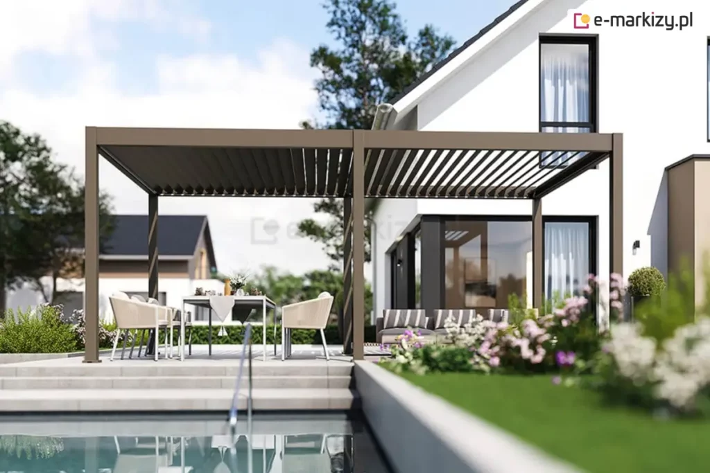 Brązowo-szara (taupe) lamelowa pergola tarasowa - model Marlo zamontowana na nowoczesnym, jasnym tarasie przy białym budynku mieszkalnym o prostej bryle. Konstrukcja pergoli opiera się na sześciu pionowych słupach o kwadratowym przekroju, wspierających ramę z ruchomymi lamelami dachowymi, które na zdjęciu są częściowo otwarte. Pod pergolą znajduje się strefa wypoczynkowa z nowoczesnym stołem jadalnianym, kremowymi krzesłami o ażurowym splocie oraz szarą sofą ogrodową umieszczoną w głębi pod zadaszeniem. Taras jest wielopoziomowy, wykonany z jasnoszarych płyt betonowych, a bezpośrednio przed nim znajduje się basen z widoczną metalową drabinką. W otoczeniu widać starannie wypielęgnowany trawnik, niską roślinność ozdobną z różowymi kwiatami oraz inne nowoczesne domy jednorodzinne w tle pod bezchmurnym, błękitnym niebem.