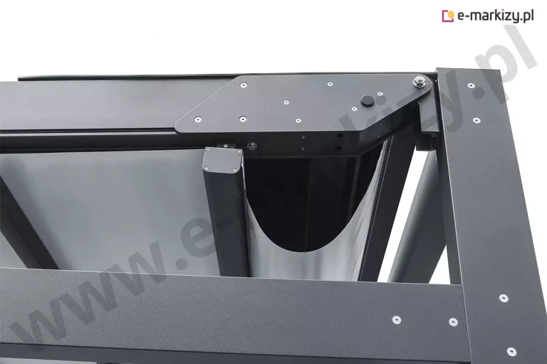 Zbliżenie na tylny węzeł konstrukcyjny pergoli Solid Aluprof, obrazujące mechanizm prowadzenia tkaniny oraz detale wykończenia aluminiowej ramy w kolorze antracytowym. Na ujęciu widoczny jest punkt styku poziomej belki obwodowej z pionowym słupem, gdzie zamontowano masywną, wielokątną płytę boczną z licznymi śrubami montażowymi, osłaniającą elementy napędowe. Pod prowadnicą znajduje się profil czołowy zadaszenia, z którego wyprowadzona jest jasna tkanina techniczna, tworząca w tym miejscu głęboką, łukowatą falę. Wnętrze konstrukcji ukazuje czarny, pionowy profil końcowy oraz system rynnowy zintegrowany z ramą, co zapewnia estetyczne ukrycie elementów funkcyjnych. Powierzchnia aluminium posiada matową, ziarnistą strukturę lakieru proszkowego, a precyzyjne spasowanie metalowych komponentów podkreśla techniczny profesjonalizm i nowoczesny design całego systemu osłonowego.