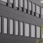 Elewacja nowoczesnego budynku biurowego wyłożona ciemnoszarymi panelami kompozytowymi o matowym wykończeniu. W pionowych, wąskich otworach okiennych zamontowano zewnętrzne rolety tekstylne typu screen w kolorze jasnoszarym. Rolety są opuszczone na różnych wysokościach, co prezentuje ich funkcjonalność w zakresie regulacji dopływu światła dziennego do wnętrz. Materiał rolet ściśle przylega do prowadnic bocznych, zapewniając estetyczny i techniczny wygląd fasady. W prawym dolnym rogu kadru widoczne są fragmenty liści młodego drzewa o zielono-żółtym zabarwieniu, kontrastujące z surową, geometryczną architekturą obiektu.