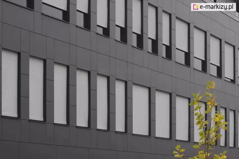 Elewacja nowoczesnego budynku biurowego wyłożona ciemnoszarymi panelami kompozytowymi o matowym wykończeniu. W pionowych, wąskich otworach okiennych zamontowano zewnętrzne rolety tekstylne typu screen w kolorze jasnoszarym. Rolety są opuszczone na różnych wysokościach, co prezentuje ich funkcjonalność w zakresie regulacji dopływu światła dziennego do wnętrz. Materiał rolet ściśle przylega do prowadnic bocznych, zapewniając estetyczny i techniczny wygląd fasady. W prawym dolnym rogu kadru widoczne są fragmenty liści młodego drzewa o zielono-żółtym zabarwieniu, kontrastujące z surową, geometryczną architekturą obiektu.