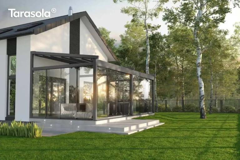 Nowoczesna, całoroczna zabudowa tarasu wykonana za pomocą pergoli Tarasola Elegancy o antracytowej konstrukcji aluminiowej, przytwierdzona do białej elewacji domu jednorodzinnego z dwuspadowym dachem. Pergola tarasowa wolnostojąca Tarasola Elegancy wyposażona jest w pełne przeszklenia boczne z przesuwnymi panelami szklanymi, które tworzą zamknięty ogród letni. Wewnątrz przestrzeni rekreacyjnej znajdują się jasnoszare, miękkie pufy oraz strefa jadalniana z krzesłami w tle. Podstawa tarasu została wykonana z gładkich, jasnych płyt betonowych, w których stopniach zamontowano punktowe, dekoracyjne oświetlenie LED. Budynek posiada nowoczesne detale, takie jak ciemne pionowe pasy na elewacji i okna połaciowe. Całość otacza rozległy, zielony trawnik oraz lasek brzozowy, a w taflach szkła widoczne są refleksy zachodzącego słońca, podkreślające transparentność i estetykę konstrukcji.