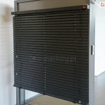 Widok na ekspozycyjny model żaluzji fasadowej C50 Slim Aluprof w kolorze antracytowym, zamontowanej w systemowej ramie aluminiowej. Zdjęcie prezentuje niemal całkowicie rozwinięty system, w którym szerokie lamele C50 są ustawione w pozycji lekko otwartej, ukazując precyzyjny układ drabinek sznurkowych. Górna rynna żaluzji jest estetycznie wykończona dopasowaną kolorystycznie kasetką, na której znajduje się subtelny, czerwony logotyp producenta. Po prawej stronie żaluzji widoczny system ręcznego sznurkowego sterowania żaluzją. Po prawej stronie profilu montażowego widoczny jest dyskretny przełącznik sterowania automatycznego, najprawdopodobniej przeznaczony do sterowania innym systemem. Ekspozycja znajduje się w jasnym, minimalistycznym wnętrzu z białymi ścianami, co pozwala na dokładną ekspozycję detali technicznych konstrukcji oraz wysokiej jakości powłoki lakierniczej elementów aluminiowych.