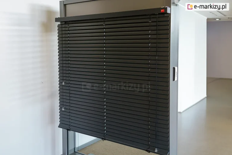 Widok na ekspozycyjny model żaluzji fasadowej C50 Slim Aluprof w kolorze antracytowym, zamontowanej w systemowej ramie aluminiowej. Zdjęcie prezentuje niemal całkowicie rozwinięty system, w którym szerokie lamele C50 są ustawione w pozycji lekko otwartej, ukazując precyzyjny układ drabinek sznurkowych. Górna rynna żaluzji jest estetycznie wykończona dopasowaną kolorystycznie kasetką, na której znajduje się subtelny, czerwony logotyp producenta. Po prawej stronie żaluzji widoczny system ręcznego sznurkowego sterowania żaluzją. Po prawej stronie profilu montażowego widoczny jest dyskretny przełącznik sterowania automatycznego, najprawdopodobniej przeznaczony do sterowania innym systemem. Ekspozycja znajduje się w jasnym, minimalistycznym wnętrzu z białymi ścianami, co pozwala na dokładną ekspozycję detali technicznych konstrukcji oraz wysokiej jakości powłoki lakierniczej elementów aluminiowych.