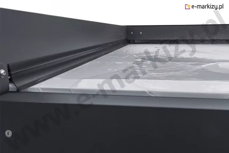 Szczegółowe zbliżenie na górną płaszczyznę zadaszenia pergoli Solid Aluprof, prezentujące właściwości wodoodporne jasnej tkaniny technicznej oraz system uszczelnień bocznych. Na powierzchni rozpiętego materiału widoczna jest warstwa wody, co demonstruje szczelność poszycia i jego zdolność do ochrony przed opadami atmosferycznymi. Wzdłuż bocznej krawędzi, przy grafitowym profilu aluminiowym, zamontowana jest czarna, elastyczna uszczelka gumowa o specjalnym profilu, która ściśle przylega do tkaniny, zapobiegając przedostawaniu się wody do wnętrza konstrukcji. Detal ten ukazuje precyzyjne dopasowanie elementów wykończeniowych, w tym widoczne śruby montażowe z łbem imbusowym osadzone w matowej, lakierowanej proszkowo ramie. Perspektywa z bliska pozwala ocenić jakość materiałów użytych do produkcji pergoli oraz techniczne rozwiązanie odprowadzania wody, które jest kluczowe dla funkcjonalności systemów tarasowych w zmiennych warunkach pogodowych.