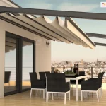 Nowoczesny dach tkaninowy rzymski Square zamontowany na przestronnym tarasie apartamentu. Konstrukcja zadaszenia opiera się na solidnych, aluminiowych profilach w kolorze antracytowym, które są przytwierdzone do elewacji budynku i wsparte na poprzecznych belkach. Jasnobeżowa, wodoodporna tkanina jest częściowo rozwinięta, tworząc charakterystyczne, głębokie fałdy podwieszone pod prowadnicami. Pod zadaszeniem znajduje się strefa jadalniana z dużym białym stołem oraz kompletem ośmiu nowoczesnych krzeseł w kolorze czarnym z białymi nogami. Taras wykończony jest jasnymi deskami kompozytowymi o wysokim połysku, w których odbijają się meble i elementy konstrukcyjne. W tle rozpościera się szeroka panorama miasta widoczna z dużej wysokości, ograniczona minimalistyczną balustradą ze stalowych linek. Na ścianie budynku, obok dużych przeszkleń, zamontowane są klasyczne kinkiety zewnętrzne, a całość oświetlona jest ciepłym światłem zachodzącego słońca, co podkreśla ekskluzywny charakter aranżacji.