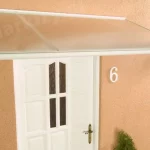 Białe drewniane drzwi z szybkami w części centralnej osłonięte zostały białym daszkiem nad drzwi Malaga z wypełnieniem z przejrzystego poliwęglanu. Drzwi i daszek kontrastują z pomarańczową elewacją. Po prawej stronie drzwi zamocowano cyfrę "6" określającą numer mieszkania, a po lewej stronie białą lampkę ze stylową obudową i widoczną żarówką.