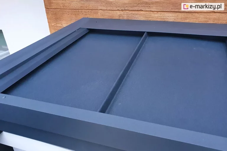 Zdjęcie przedstawia zbliżenie na górny narożnik aluminiowej konstrukcji pergoli tarasowej Luxury w kolorze ciemnego antracytu. Głównym elementem widocznym na fotografii jest systemowy daszek ochronny wykonany z gładkich paneli, który jest osadzony wewnątrz ramy konstrukcyjnej. Daszek ma za zadanie zabezpieczać zwiniętą tkaninę zadaszenia przed czynnikami atmosferycznymi. Panele są precyzyjnie spasowane i oddzielone od siebie cienkimi przetłoczeniami wzmacniającymi. W górnej części kadru widoczny jest fragment elewacji budynku wykończony deską elewacyjną w naturalnym, miodowym kolorze drewna z wyraźnym rysunkiem słojów. Po lewej stronie widać fragment białej ściany budynku, co tworzy kontrast z ciemną ramą pergoli. Perspektywa z góry pozwala dokładnie ocenić matowe wykończenie lakieru proszkowego na profilach aluminiowych oraz stabilność montażu elementów osłonowych.