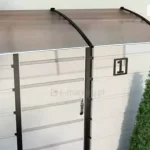 Zdjęcie przedstawia nowoczesny daszek naddrzwiowy o konstrukcji modułowej, zamontowany na jasnej elewacji budynku o poziomym układzie paneli. Zadaszenie składa się z dwóch łukowatych segmentów wypełnionych półprzezroczystymi płytami z poliwęglanu komorowego w kolorze dymionego brązu. Płyty są osadzone w wytrzymałych, czarnych wspornikach wykonanych z tworzywa lub aluminium, które mają profilowany kształt i są przymocowane bezpośrednio do ściany. Konstrukcja jest wzmocniona listwami wykończeniowymi na krawędziach, zapewniającymi sztywność i odprowadzanie wody. Daszek osłania wejście do budynku, przy którym widoczny jest pionowy fragment ciemnej framugi oraz tabliczka z numerem posesji jeden. W otoczeniu znajdują się trzy dekoracyjne, stożkowate krzewy iglaste o żywozielonym kolorze, ustawione symetrycznie po obu stronach wejścia.