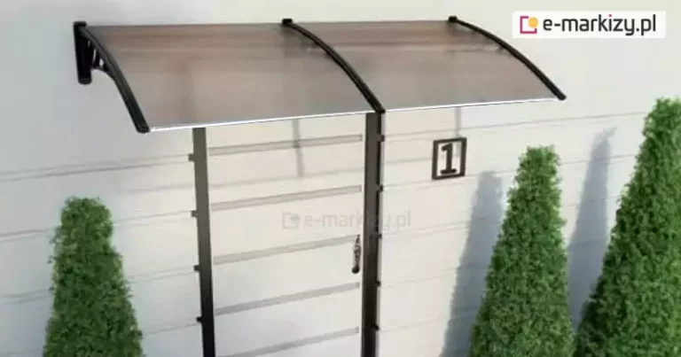 Zdjęcie przedstawia nowoczesny daszek naddrzwiowy o konstrukcji modułowej, zamontowany na jasnej elewacji budynku o poziomym układzie paneli. Zadaszenie składa się z dwóch łukowatych segmentów wypełnionych półprzezroczystymi płytami z poliwęglanu komorowego w kolorze dymionego brązu. Płyty są osadzone w wytrzymałych, czarnych wspornikach wykonanych z tworzywa lub aluminium, które mają profilowany kształt i są przymocowane bezpośrednio do ściany. Konstrukcja jest wzmocniona listwami wykończeniowymi na krawędziach, zapewniającymi sztywność i odprowadzanie wody. Daszek osłania wejście do budynku, przy którym widoczny jest pionowy fragment ciemnej framugi oraz tabliczka z numerem posesji jeden. W otoczeniu znajdują się trzy dekoracyjne, stożkowate krzewy iglaste o żywozielonym kolorze, ustawione symetrycznie po obu stronach wejścia.