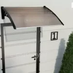 Grafika przedstawia wizualizację pojedynczego daszka drzwiowego model Coppoline, zamontowanego na białej, poziomo boniowanej elewacji nad jasnymi drzwiami wejściowymi. Konstrukcja zadaszenia składa się z dwóch czarnych wsporników o ozdobnym, ażurowym wypełnieniu z geometrycznymi motywami, które podtrzymują lekko nachyloną połaci wykonaną z poliwęglanu w odcieniu dymnego brązu. Płyta jest ujęta w ciemne profile boczne, a jej krawędź czołowa wykończona jest estetyczną listwą. Obok drzwi widoczna jest tabliczka z numerem posesji jeden, a całą aranżację uzupełniają symetrycznie ustawione przed budynkiem dwa wysokie, formowane krzewy iglaste w kształcie stożków. Daszek charakteryzuje się prostym, nowoczesnym designem, zapewniając ochronę strefy wejścia przed opadami atmosferycznymi przy zachowaniu dużej przepuszczalności światła.
