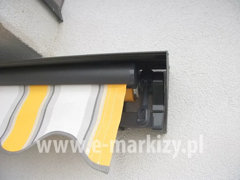 Prawy bok markizy bez kasety z opcjonalnym daszkiem do markiz, całość w kolorze srebrzysty grafit, z poszyciem w białe, szare i żółte pasy. Markiza z napędem ręcznym i falbaną falowaną zawieszona pod balkonem na ścianie z białym tynkiem strukturalnym