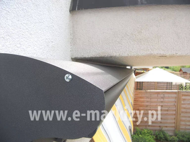 Zamocowany tuz pod płytą balkonu daszek do markizy w kolorze srebrzysty grafit z widocznym boczkiem skręconym z blachą górną za pomocą srebrnej śruby, w tle widoczny płot z drewnianych paneli oraz pawilon z białym dachem
